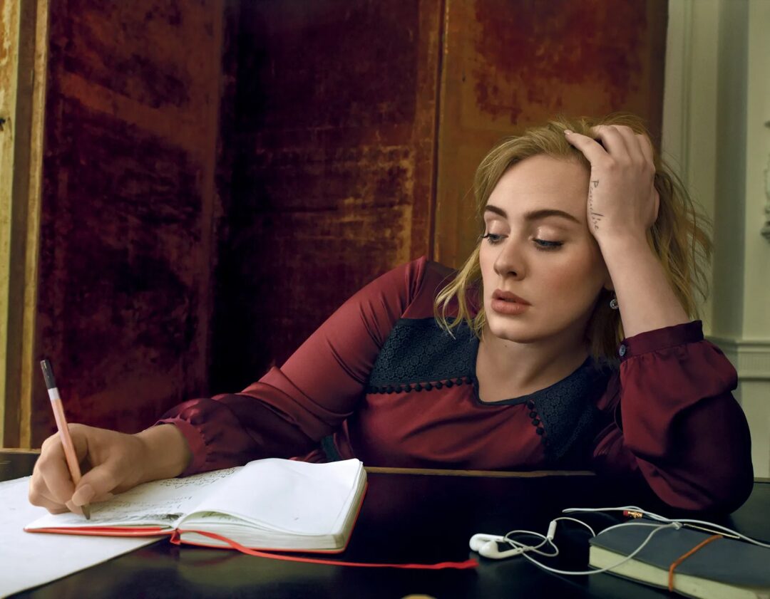 Adele writing in a notebook with a pencil and earphones on the table, Burberry dress